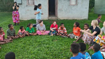 A group of children and adults sit in a circle on grass outside a concrete building during a Fiji school trip, with one person standing and another taking a photo in the background.