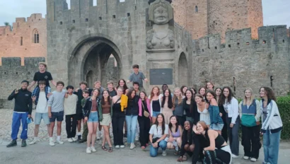 A group of young people on a France school trip pose for a photo in front of a stone castle entrance, featuring an arched doorway and a large stone bust.