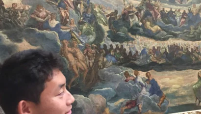 A student on a France school trip, gazes at a large, detailed Renaissance painting depicting a crowded, heavenly scene filled with figures in flowing robes.