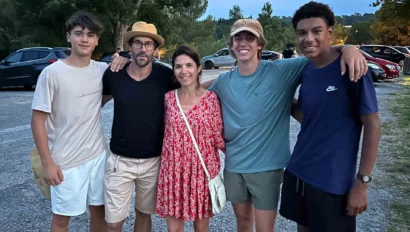 Five people stand together outdoors in a parking lot, posing for a group photo during their France school trip; trees and parked cars are visible in the background.