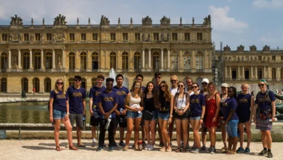 A group of young people on a France school trip pose in front of the Palace of Versailles, with some wearing matching purple shirts, on a sunny day.