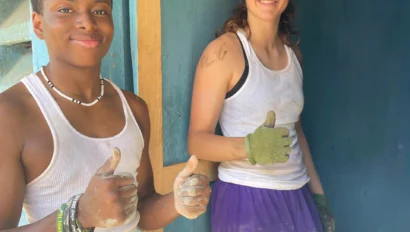 Two people wearing work clothes and giving thumbs up, standing by a blue wooden wall with paint and dirt on their clothes and hands after a successful Dominican Republic school trip project.