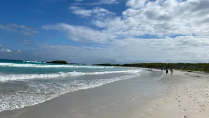 Sandy beach with gentle waves under a partly cloudy sky. A few people walk along the shoreline, and footprints are visible in the sand.
