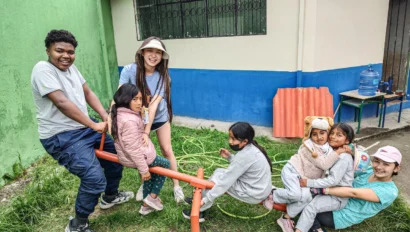 Seven children and a woman enjoy an orange seesaw in a grassy outdoor area near a building with green and blue walls during an Ecuador school trip.