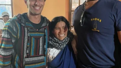Three people stand indoors smiling for a photo during an Ecuador school trip. Two young men are on either side of a woman in traditional clothing, with tan walls and windows visible in the background.
