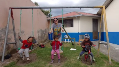 During an Ecuador school trip, an adult pushes two children on swings while another child sits on a bicycle in a grassy yard with a painted wall and building in the background.