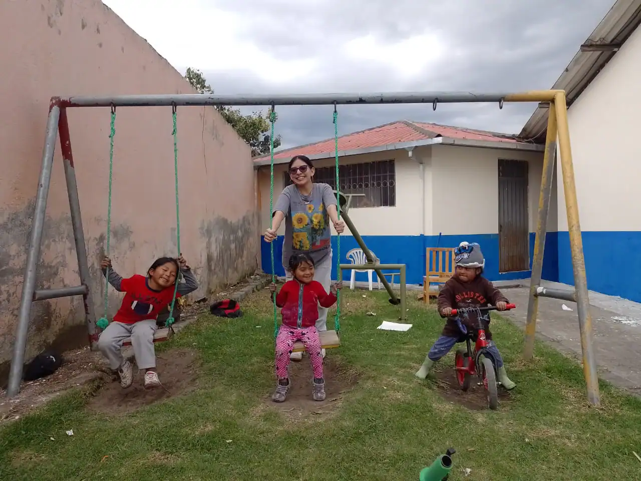 During a school trip to Ecuador, an adult pushes two children on swings while a third child sits on a red bicycle in a grassy yard beside a building with a blue wall.