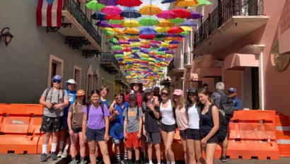 A group of people stand on a cobblestone street under a canopy of colorful umbrellas. Orange barricades line the sides. A Puerto Rican flag is visible on a building.