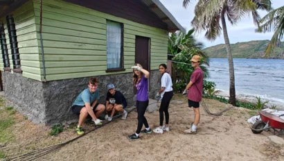 A group of people stand beside a green house near the ocean. They appear to be involved in a landscaping or construction project, with a wheelbarrow and tools nearby.