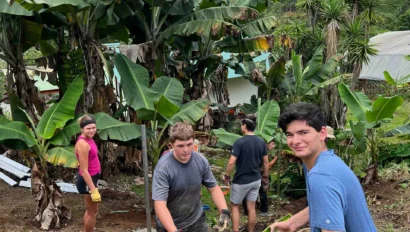 Four people work together outdoors, carrying a bucket and handling soil or plants near banana trees, with green hills and buildings in the background—a perfect snapshot of service trips for teens.