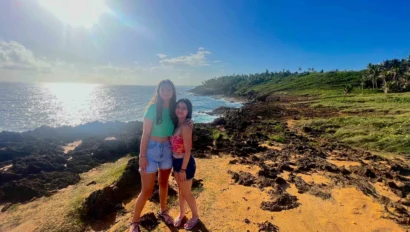 Two people stand on rocky terrain by the ocean under a clear blue sky, with sunlight reflecting on the water.