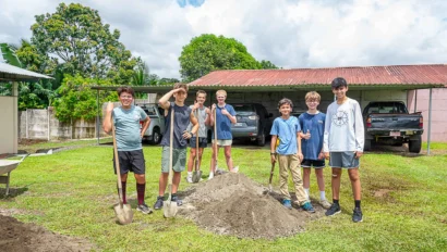 A group of young people standing on grass with shovels, next to a pile of dirt. Cars are parked in the background.