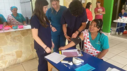 Three healthcare workers take a seated woman's blood pressure at a health clinic. Papers and a blood pressure monitor are on the table. Other people are in the background.