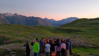 A group of people gathered around a campfire in a grassy mountainous landscape at sunset.