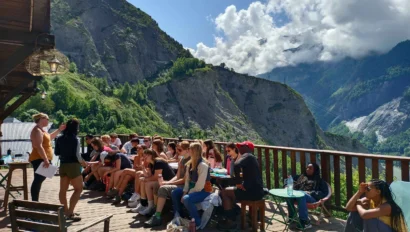 A group of people sitting and standing on a wooden deck overlooking a mountainous landscape under a partly cloudy sky.