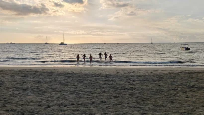 Silhouettes of people jumping in waves at a sandy beach during sunset, with sailboats in the background.