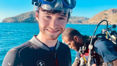 A person in a wetsuit and snorkel mask gives two thumbs up on a boat during their fiji summer service trip, with another person in scuba gear in the background and blue water and hills visible.