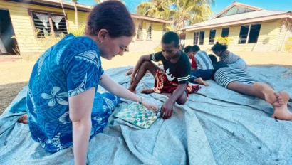 A student is teaching several children as they sit on a tarp outdoors, playing a board game together, with houses and palm trees visible in the background.