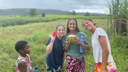 Four people smiling in a green field, two holding fruit. Overcast sky above.