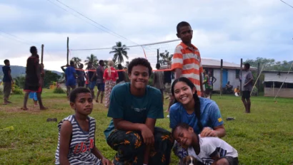 A group of children and a woman sit and kneel on grass during a summer service trip, watching a volleyball game with tropical trees and houses in the background.