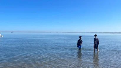 Two boys stand in shallow water near the shore under a clear blue sky, enjoying the calm sea and distant boats during their summer service trip.