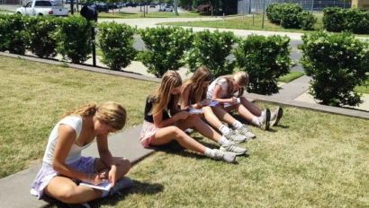 Five girls sit on the grass outside on a sunny day, each writing or drawing in notebooks, as part of a travel writing program, with a road, bushes, and stadium lights in the background.