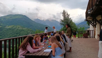 A group of people sit at wooden tables on a deck overlooking green mountains, brainstorming for their travel writing program, while others stand nearby under a cloudy sky.