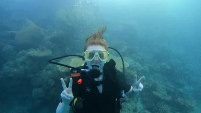 A scuba diver with red hair, wearing yellow goggles, holds up peace signs underwater with coral in the background.