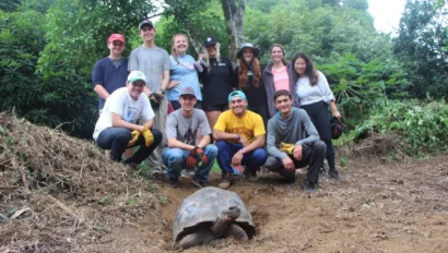 A group of ten people wearing casual clothes stands behind a large tortoise in a lush, green outdoor setting.
