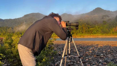 Person in black jacket and boots uses a spotting scope on a tripod in a rocky field, with green hills and blue sky in the background.