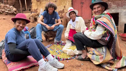 Four people in a rural setting are seated on a blanket, surrounded by colorful textiles and corn. They are wearing casual and traditional clothing, with a rustic building in the background.