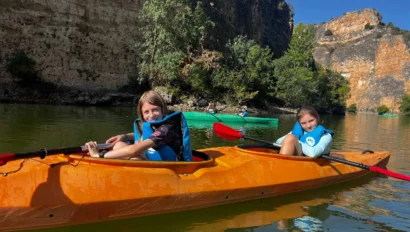 Two kids in life vests paddle an orange kayak on a calm river, surrounded by rocky cliffs and greenery under a clear blue sky.