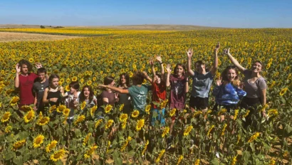 A group of people stands in a sunflower field under a clear blue sky, waving.