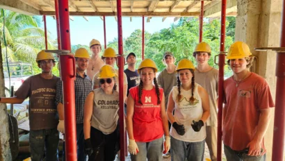 A group of people wearing yellow hard hats and casual clothes pose together at a construction site, showcasing their service travel for teens, with greenery visible in the background.