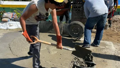 Workers in hard hats and gloves mix and pour concrete on a construction site under a clear blue sky.