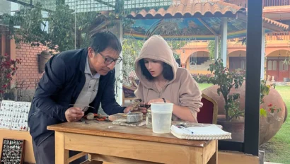 Two people focusing on a small object at a wooden table in a bright room with large windows overlooking a garden. A gas cylinder is nearby.