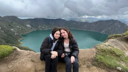 Two people sitting on a stone ledge with a scenic backdrop of a large blue lake surrounded by hills and cloudy skies.