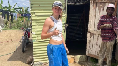 A person in a white tank top and blue shorts stands outside a corrugated metal building, giving a thumbs up during a service travel for teens trip, with several buckets on the ground and another person nearby.