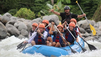 A group of people wearing helmets and life jackets paddle a blue inflatable raft through whitewater rapids, with a guide steering at the back—an exciting service travel for teens adventure.