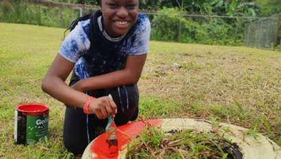A person smiles while painting the edge of a white tire planter orange in an outdoor grassy area during a Costa Rica school trip, with a paint can nearby.
