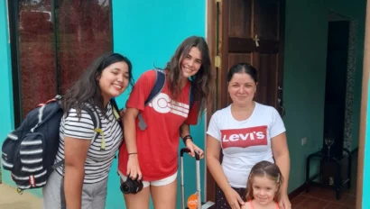 Three women and a girl stand outside a turquoise building. The women have luggage and backpacks, and they are smiling.