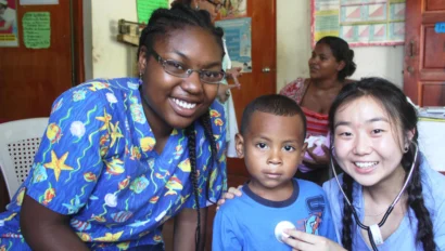 Two women and a young boy pose and smile in a brightly lit clinic room, likely part of a medical study abroad program; one woman places a sticker on the boy’s shirt while others are seen in the background.