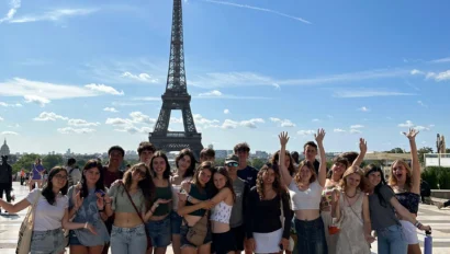 A group of people on a student trip to France poses and smiles in front of the Eiffel Tower on a sunny day with a clear blue sky.