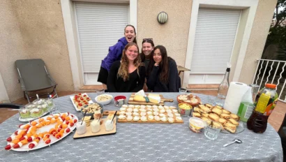 Four people pose behind a table with various snacks, including skewers, cheese, and pastries, outside a building with closed shutters.