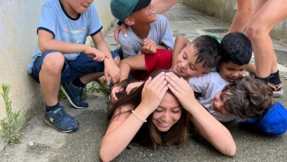 A group of kids and a young woman are playing on the ground outside, with the kids piled on top of the woman—perhaps reminiscing during a joyful student trip to France—as she lies on the pavement, smiling.