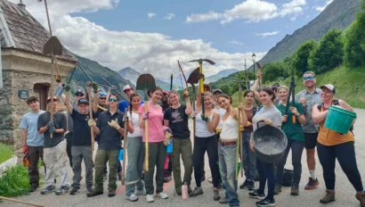 A group of people on a France student trip stand outdoors on a road in a mountainous area, holding various tools and buckets, posing for the camera.