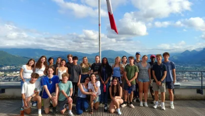 A group of young people stands together on a balcony with a French flag, overlooking a mountainous landscape. The sky is partly cloudy.