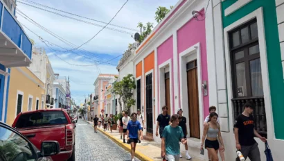 People walk along a colorful street with vibrant buildings in a historic district. Cars are parked along the cobblestone road.
