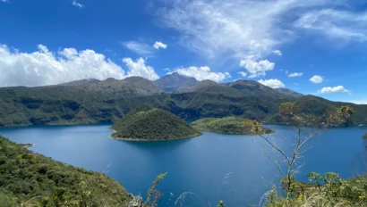 A scenic view of a blue lake surrounded by green hills and mountains under a blue sky with scattered clouds.