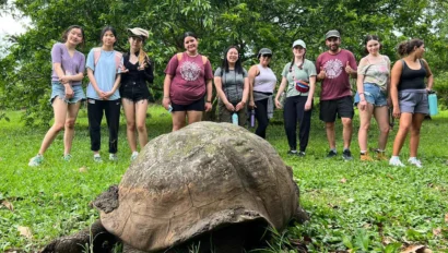 A group of people stands in a grassy area behind a large tortoise, enjoying summer travel with trees in the background.